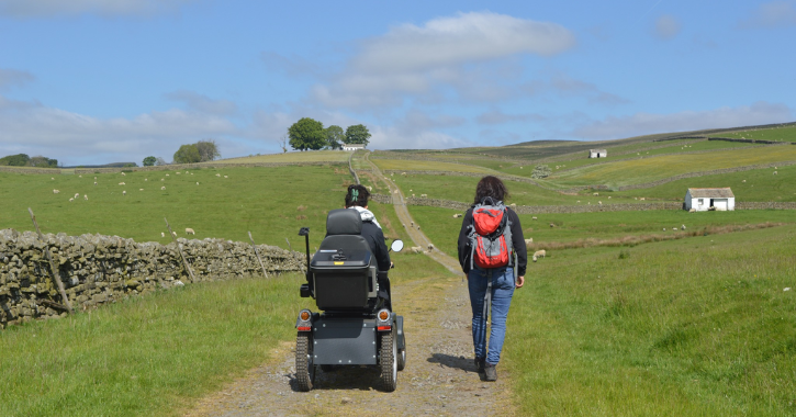 Woman walking next to a person in a tramper mobility scooter surrounded by the Durham Dales countryside.